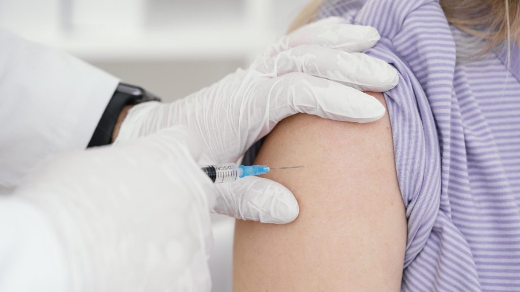 z lewej strony ręka w białej rękawiczce, z prawej ramie, moment podania szczepionki