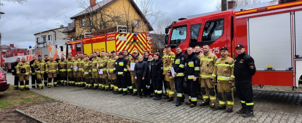 Na zdjęciu znajdują się druhowie ochotnicy ustawieni w dwuszeregu, którzy brali udział w ćwiczeniach bojowych. W tle znajdują się pojazdy strażackie.