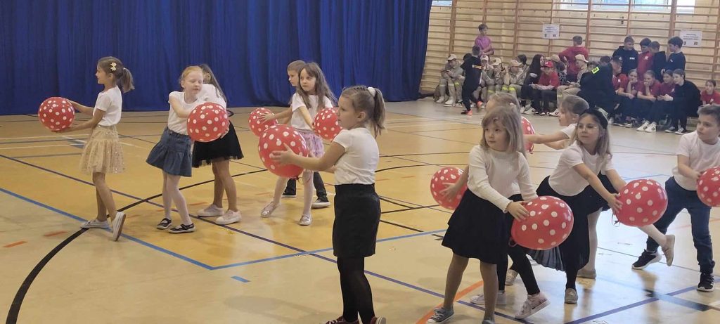 Na obrazku jest grupa dzieci trzymająca czerwone balony w sali gimnastycznej. Dzieci wyglądają na szczęśliwe i podekscytowane, a sala jest jasna i przestronna.
