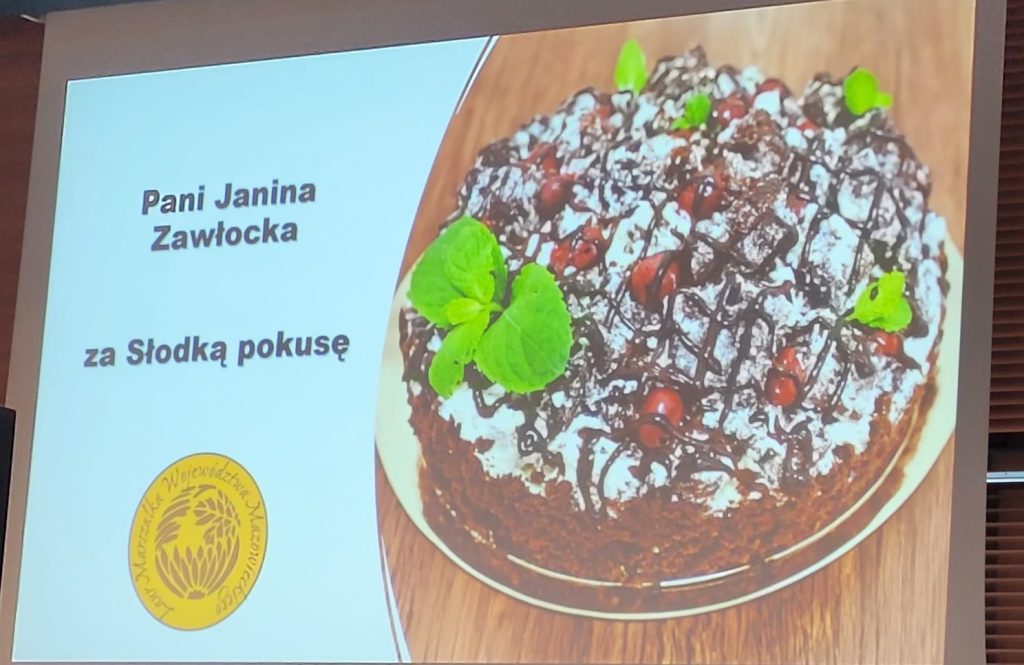 Na slajdzie prezentacyjnym jest pyszne ciasto czekoladowe udekorowane zielonemi składnikami i jagodami, co może sugerować kontekst kontekstu czy konkursu prania. Deser wygląda apetycznie, a jego estetyczna prezentacja stosowana na wysokim poziomie umiejętności cukierniczych.