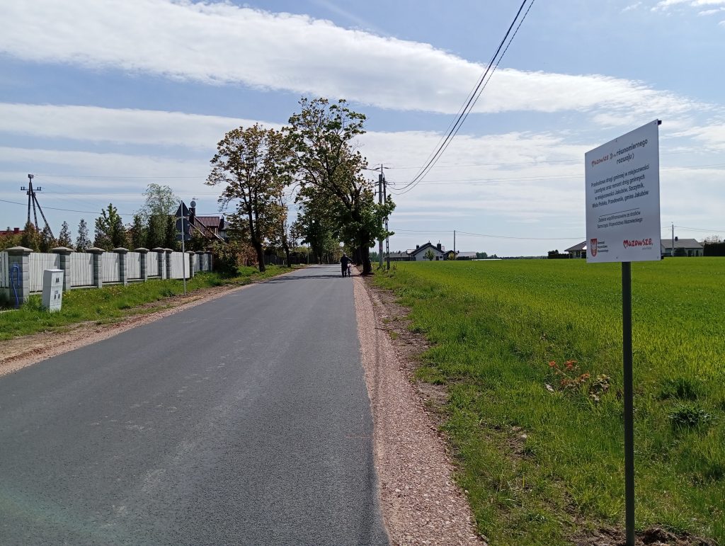 Droga asfaltowa prowadzi przez wiejski krajobraz z zielonym polem po prawej. Po lewej jest ogrodzenie i kilka domów. Na pierwszym planie stoi biały znak informacyjny.