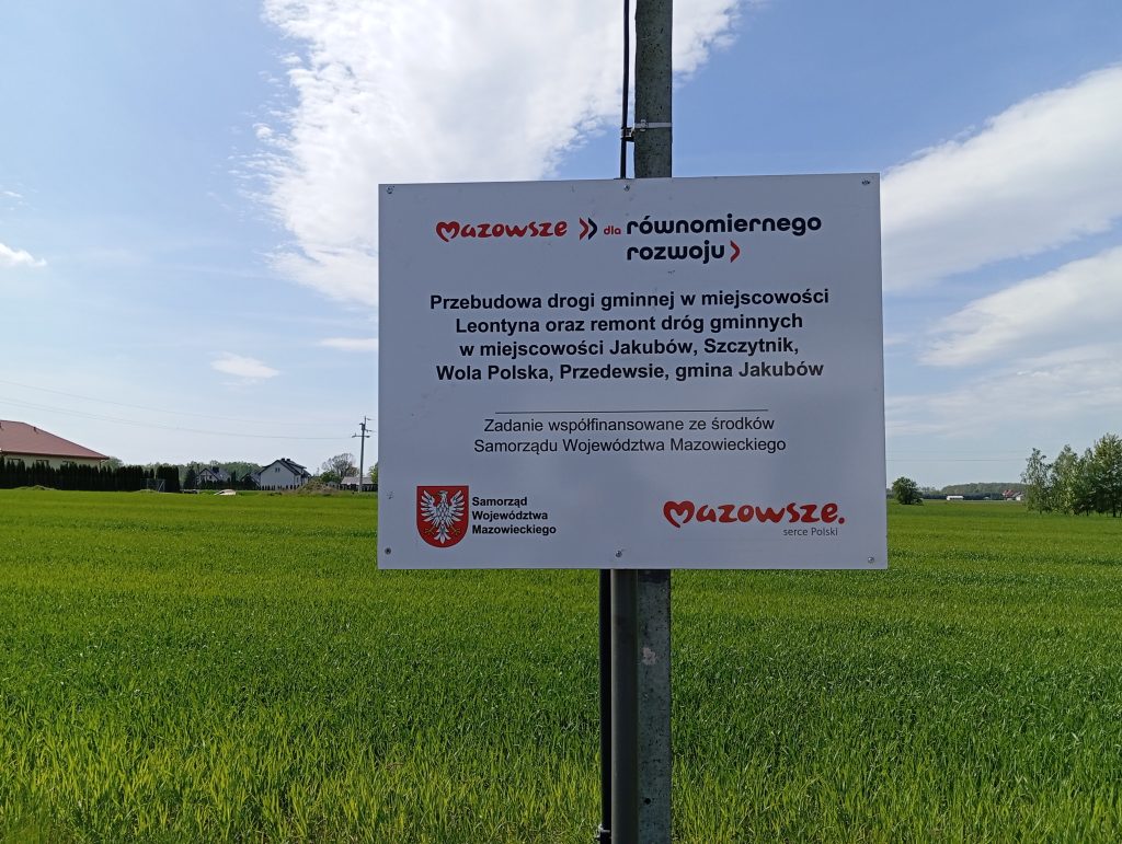 Tablica informacyjna na tle zielonego pola i niebieskiego nieba. Zawiera informacje o projekcie drogowym w gminie Jakubów. Widoczne logo Mazowsza.