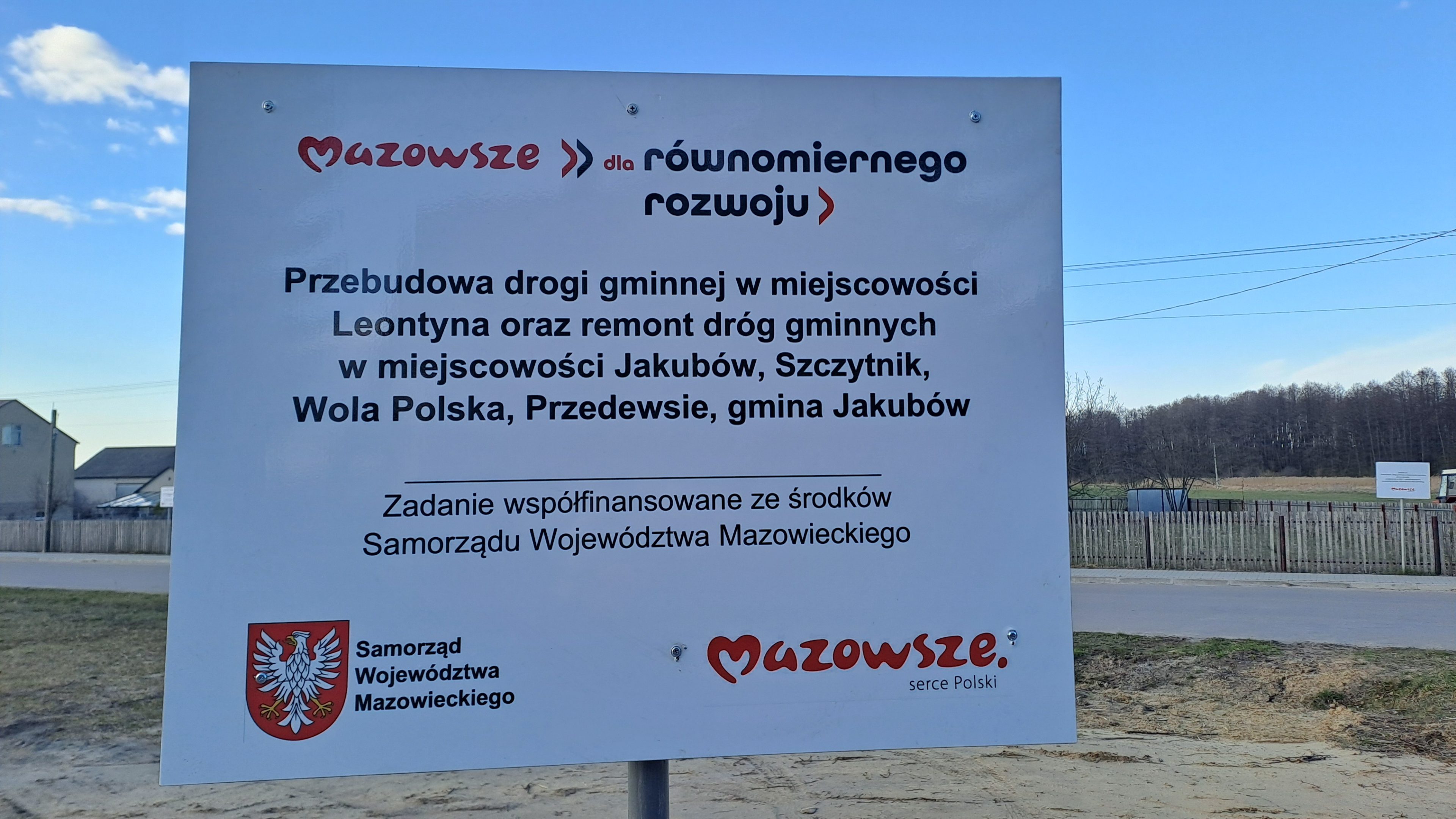 Na obrazku widać znak informacyjny projektu budowy dróg w kilku polskich miastach, finansowanego przez rząd województwa mazowieckiego. Znak zawiera logo i informacje o instytucjach oraz szczegóły dotyczące lokalizacji i rodzaju znalezionych prac drogowych.
