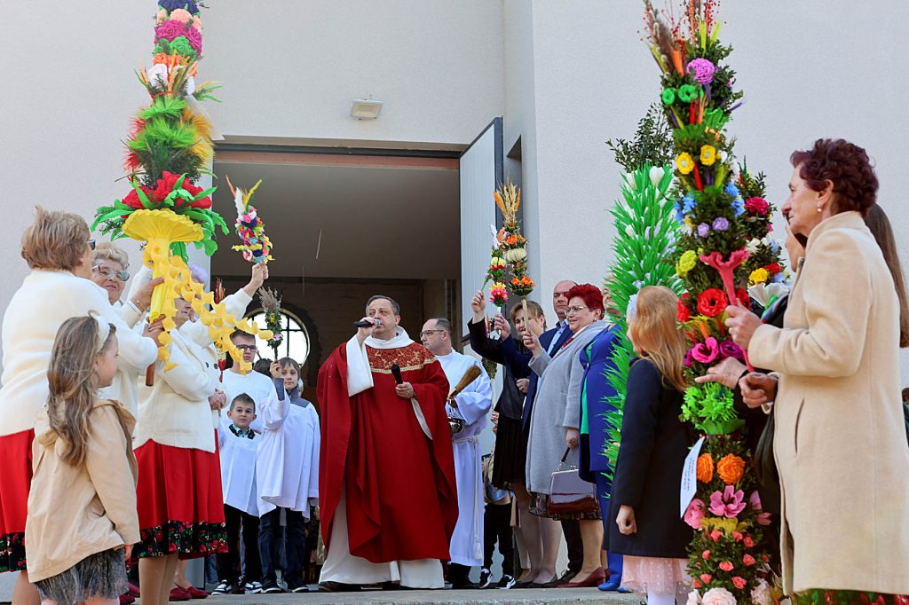 Na obrazku jest proces religijny, w którym centralna jest mężczyzna ubrany w wolną szatę. Wokół niego znajdują się osoby trzymające wysokie, barwne dekoracje kwiatowe, wszyscy zgromadzeni przed wejściem do kościoła.