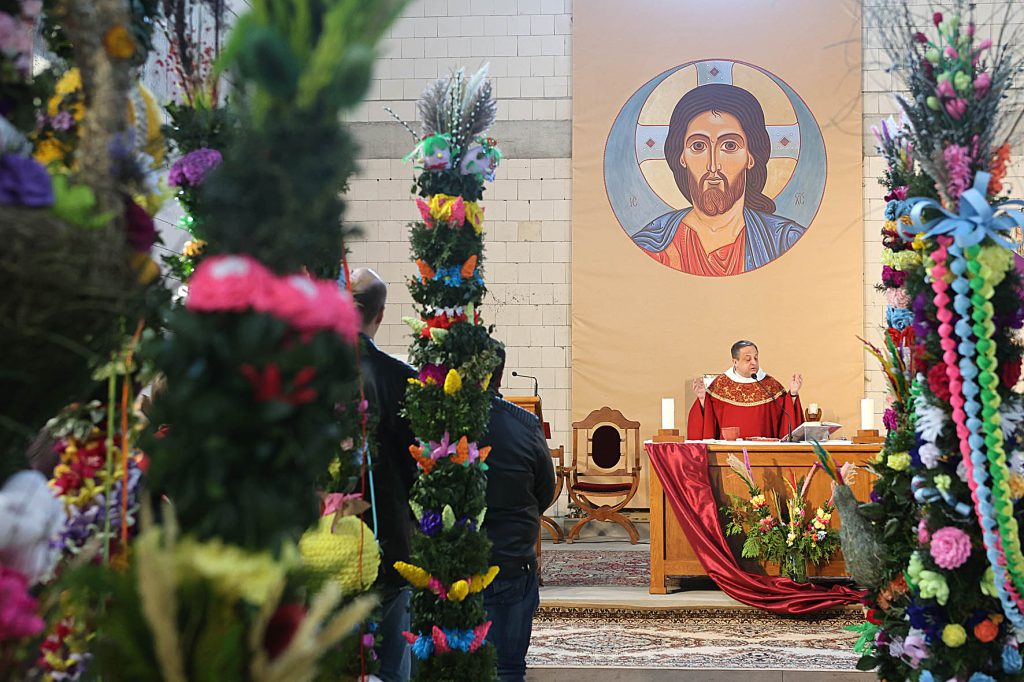 Na obrazie ksiądz ubrany w czerwone szaty liturgiczne przemawia przy ołtarzu, nad którym widnieje duża ikona religijna. Cała scena otoczona jest elementami roślinnymi, które dodają uroczystego charakteru.