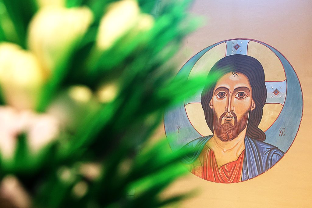 Na obrazku znajduje się ikona przedstawiająca postać religijną z aureolą, udostępnioną w tle. Na pierwszym planie, częściowo zasłaniając, widać rozmyte zielone i białe kwiaty.