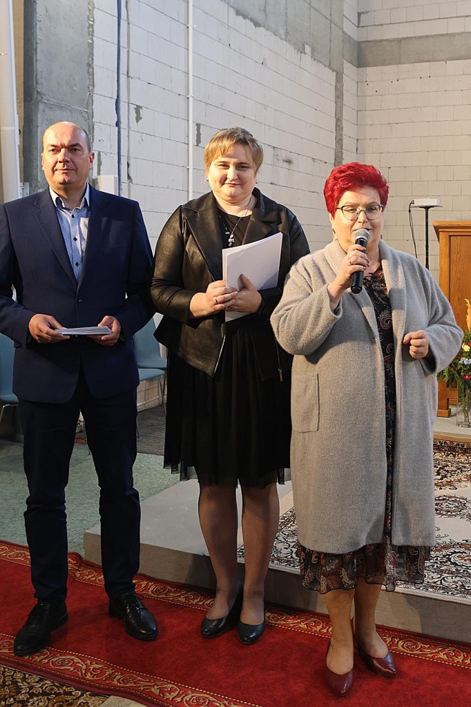 Na rynku znajdują się trójka osób stojących na niewielkiej płycie. Kobieta z rudymi włosami mówi do mikrofonu, podczas gdy pozostałe dwie osoby trzymają kartki papieru, a całość odbywa się w pomieszczeniu z szarymi ceglastymi ścianami i czerwonym dywanem.