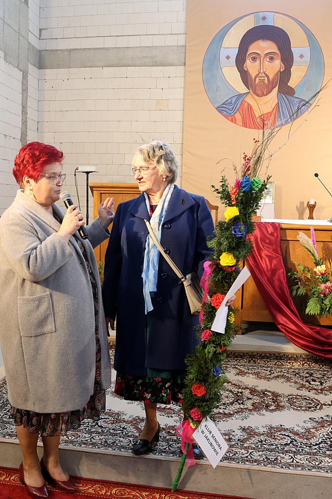 Na obrazku znajdują się dwie kobiety, które są obok kwiatowej i mikrofonu w kościelnym otoczeniu. W tle jest duża ikona religijna, która tworzy kontekst duchowy poszczególnych scen.