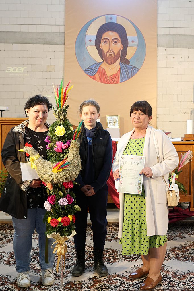 Na obrazku znajdują się trzy osoby znajdujące się wewnątrz kościoła, utrzymujące ozdobną kompozycję kwiatową w obudowie koguta. Na ścianie za nimi jest ikona religijna.