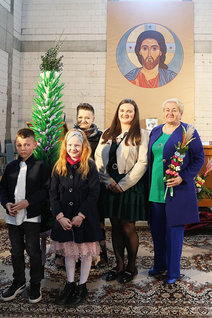 Na obrazku pięć osób stojących w pomieszczeniu, pozujących na tle ikon religijnych. Na przodzie dwóch dzieci, za które znajdują się trzy elementy klasyczne, a po lewej stronie jest zainstalowana instalacja.
