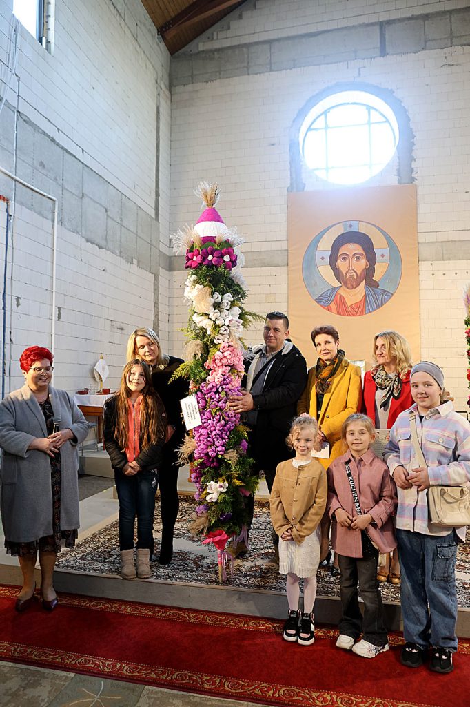 Na miejscu znajduje się grupa osób stojących w pomieszczeniu obok wysokiej częstotliwości kwiatowej. W tle widać, że przedstawia postać religijną, co podkreśla sakralny charakter otoczenia.