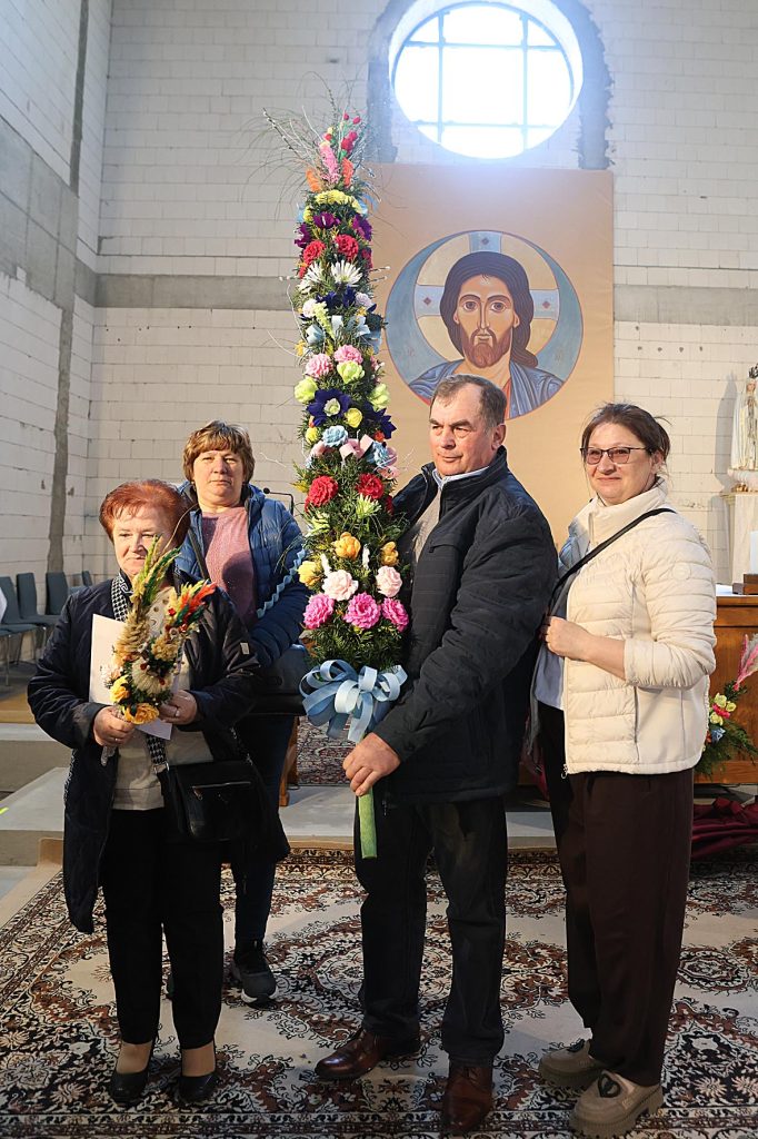 Na obrazku znajdują się cztery osoby w pomieszczeniu, z czego trzymają się wysokie i kolorowe bukiety kwiatowe. W tle jest ikona religijna, co może sugerować, że zawiera ona charakter uroczysty lub religijny.