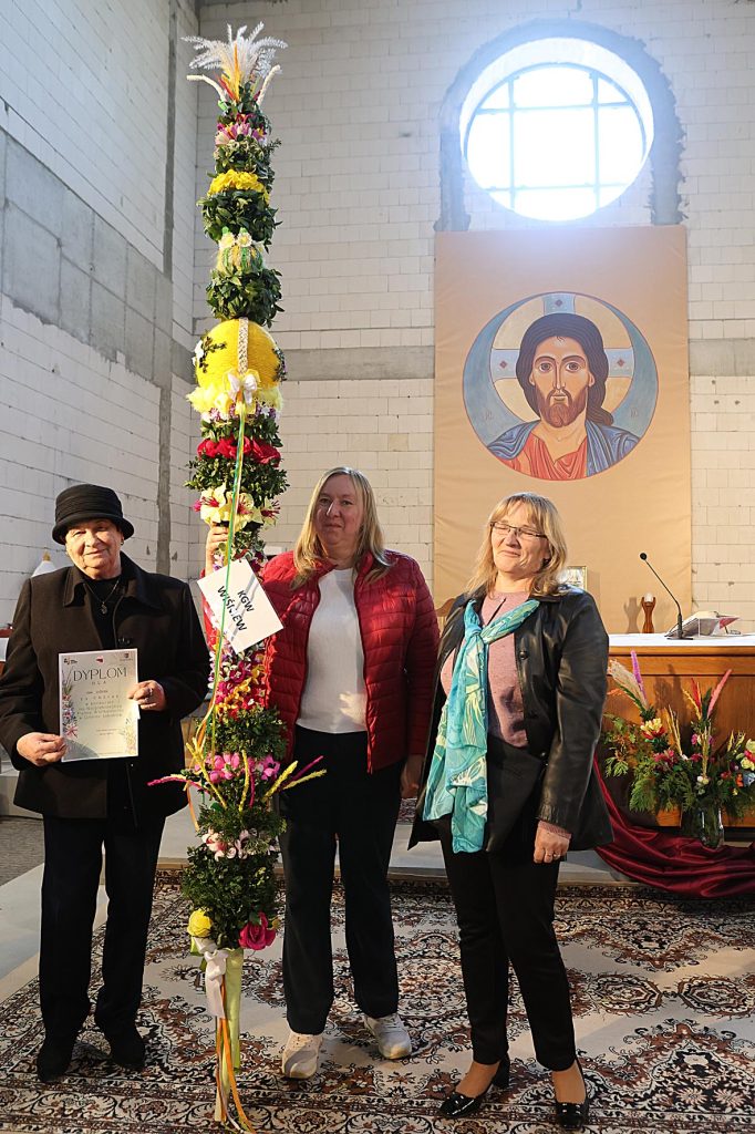 Na obrazku znajdują się trzy osoby, które są umieszczone w obok, wyposażonego w strukturę kwiatowego. W tle widoczny jest obraz przedstawiający postać religijną, a osoba po lewej stronie trzyma dyplom.