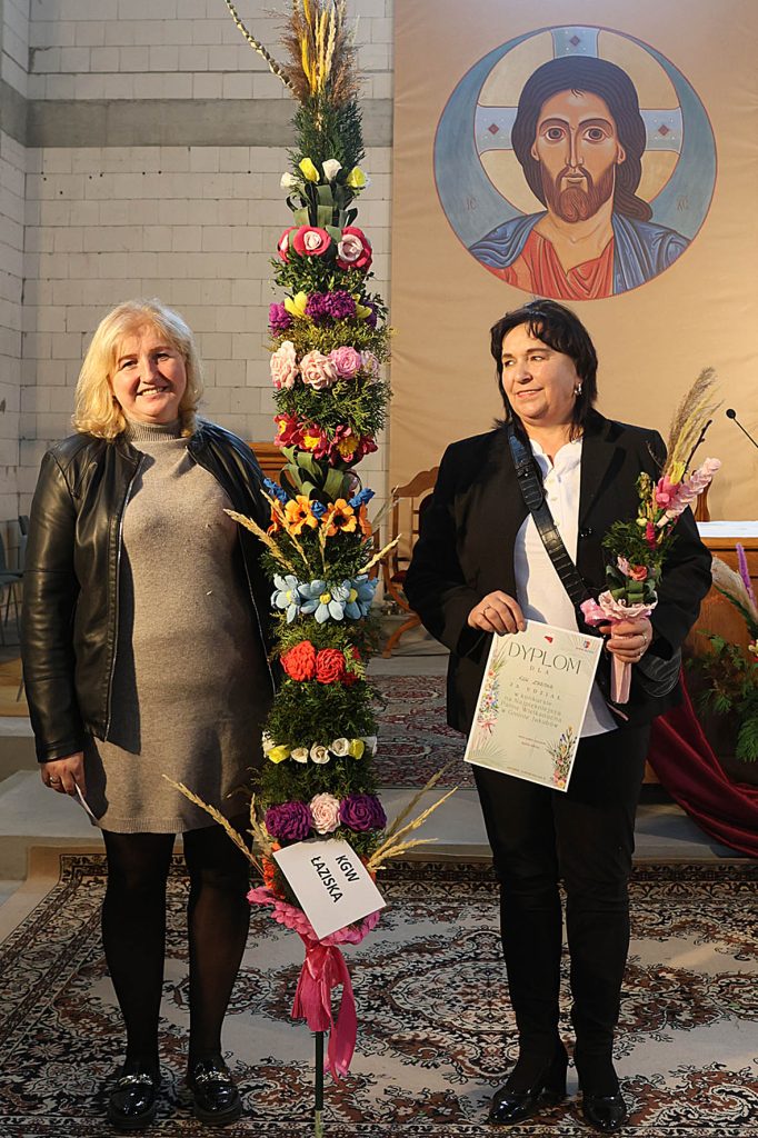 Na dwie osoby występujące wewnątrz budynku, posiadające wysokie aranżacje kwiatowe oraz certyfikat. Za nimi, na ścianie, jest duża ikona religijna.