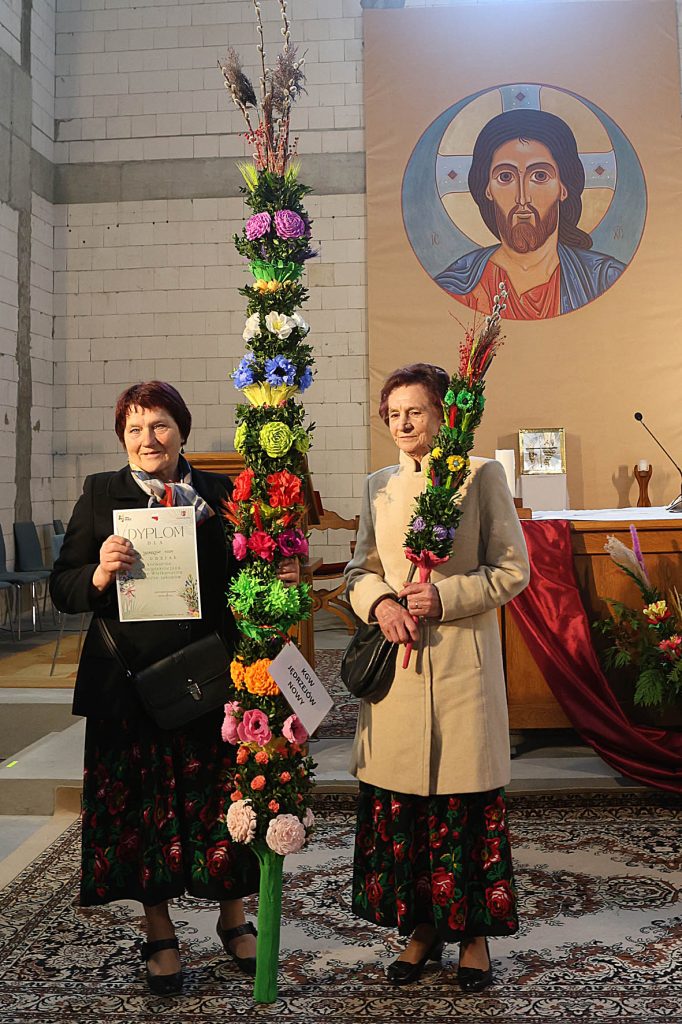 Na obrazku znajdują się dwie kobiety, które są w kościele, trzymające wysokie, kolorowe kwiaty kwiatowe. W tle jest ikona religijna.