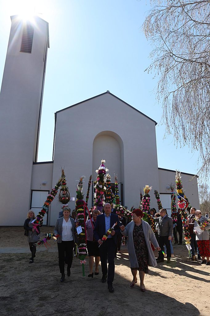 Na obrazku widać grupę osób niosących kwiaty, przemieszczające się przed białym kościołem. Tło stanowi bezchmurne niebo, które upamiętnia uroczysty charakter wydarzeń.