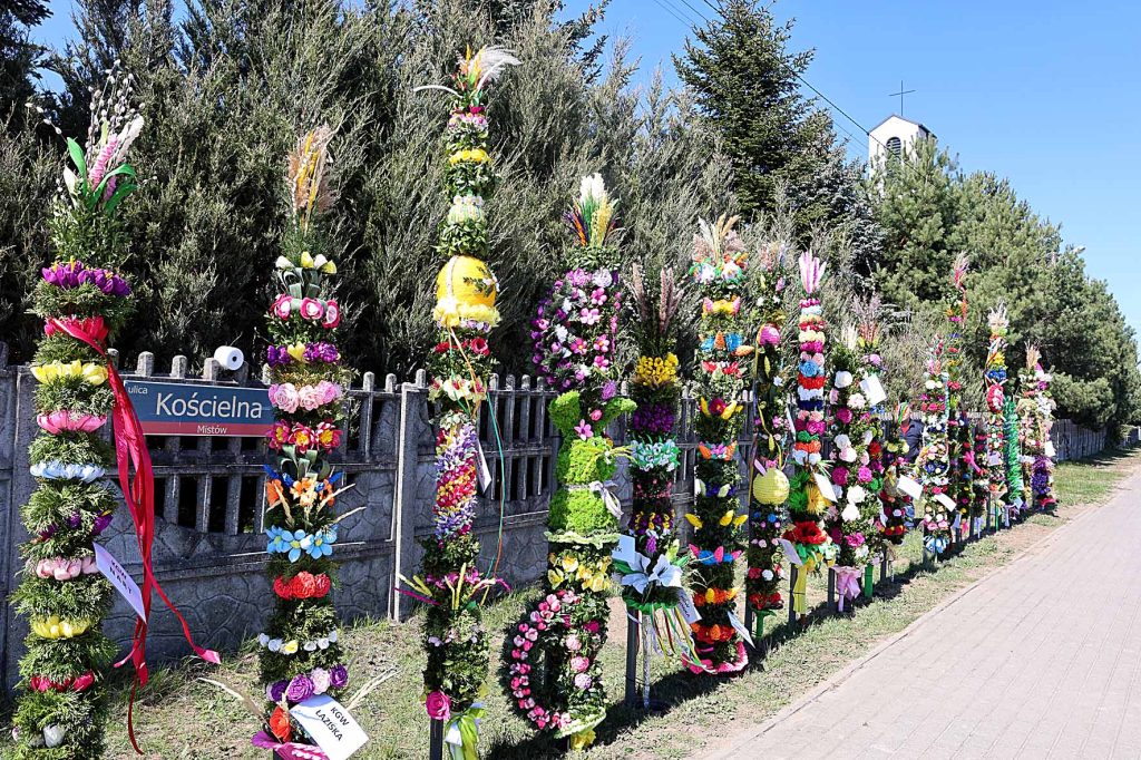 Na obrazku znajdującym się kolorowe, wysokie palmy wielkanocne, rozwiązanie obok drewnianego ogrodzenia, a w tle są drzewa. Obok znajduje się mały znak z napisem 
