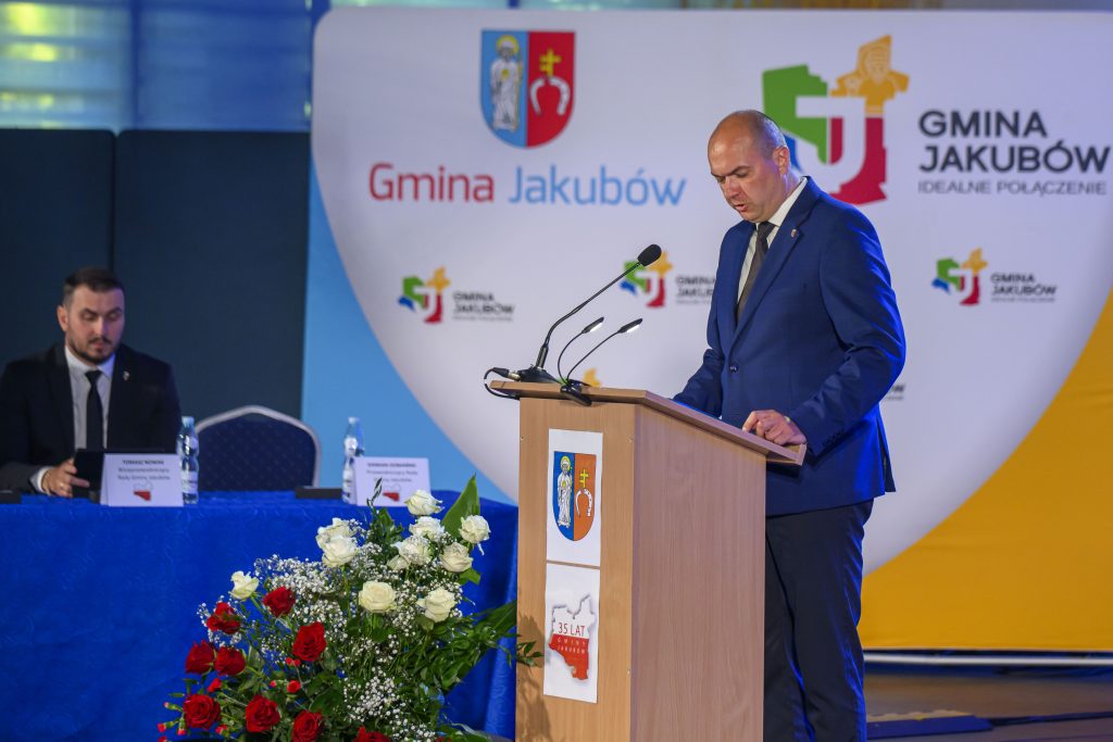 Mężczyzna w garniturze przemawia przy drewnianej mównicy, obok siedzi drugi mężczyzna. Tło jest białe z napisem Gmina Jakubów, a na pierwszym planie widać kompozycję z biało-czerwonych kwiatów.