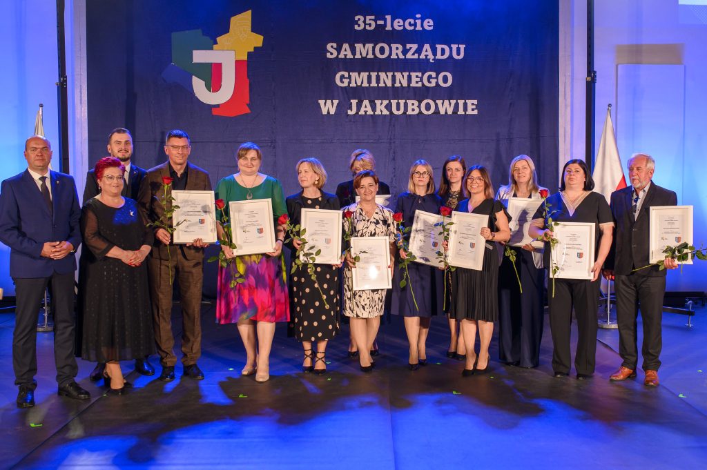 Grupa osób na scenie z dyplomami i różami na tle niebieskiego baneru. Baner informuje o jubileuszu 35-lecia samorządu w Jakubowie. Większość ubrana elegancko, tworzy formalną atmosferę wydarzenia.