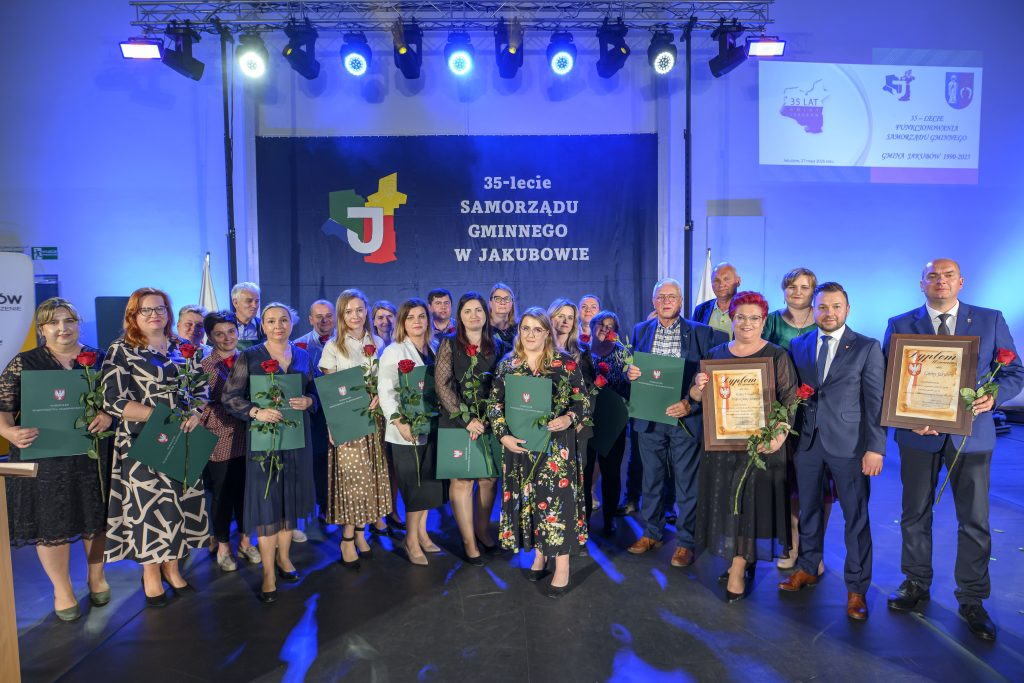 Grupa ludzi stoi na scenie z zielonymi teczkami i różami. Tło jest niebieskie z napisem o 35-leciu samorządu. Oświetlenie jasne, dominują kolory granatowy i zielony.