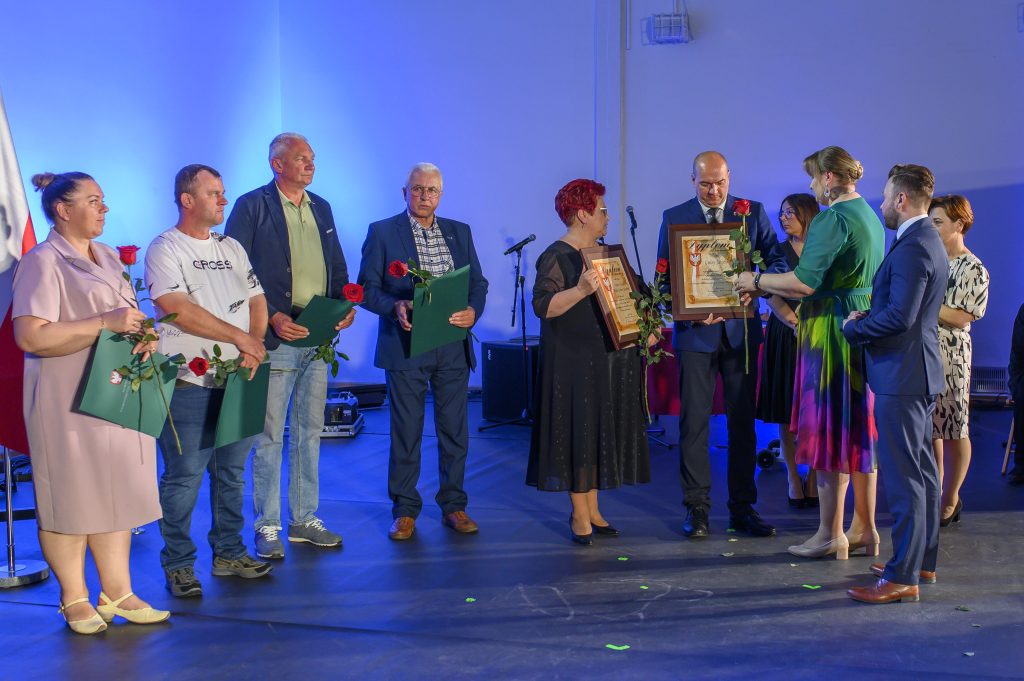 Na zdjęciu jest grupa osób na scenie w świetle niebieskich reflektorów. Każda trzyma dyplomy i czerwone róże. Tło jest jednolite, niebieskie.
