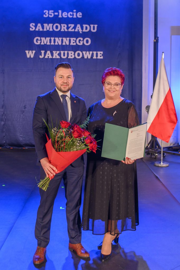 Dwójka ludzi stoi obok siebie; mężczyzna trzyma bukiet czerwonych róż, kobieta dokument. W tle granatowa kurtyna z napisem i polska flaga.