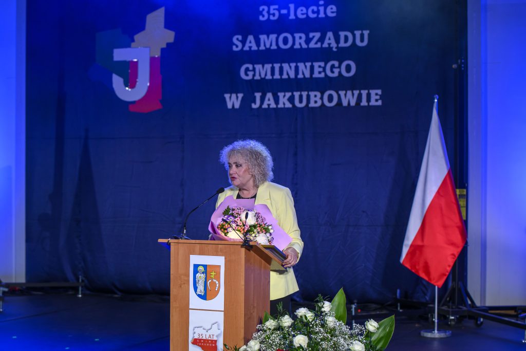Kobieta przy mównicy z bukietem kwiatów, na niebieskim tle z napisem 35-lecie Samorządu Gminnego w Jakubowie, obok flaga Polski.