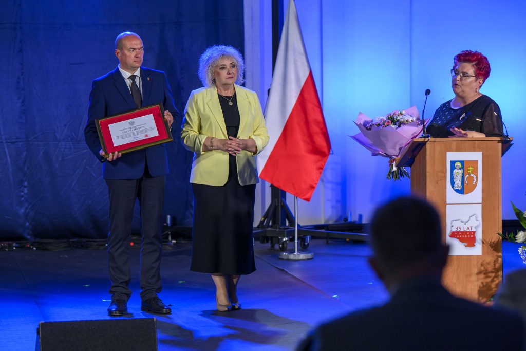 Mężczyzna i kobieta stoją na scenie z certyfikatem, w tle biało-czerwona flaga. Obok kobieta przemawia przy mównicy z bukietem kwiatów. Ściany są niebiesko oświetlone.
