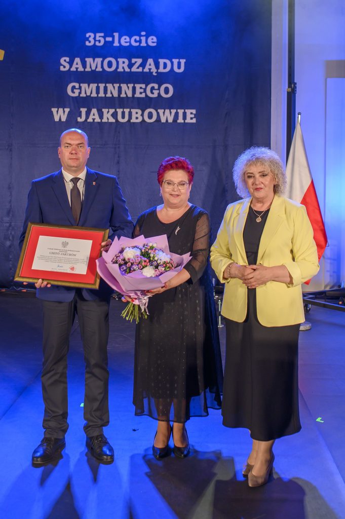 Trzy osoby pozują z dyplomem i bukietem na tle ciemnoniebieskiej kurtyny. Tło: napis 35-lecie Samorządu Gminnego. Po prawej widoczna polska flaga.