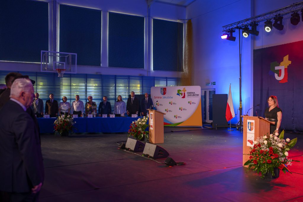 Na zdjęciu jest uroczystość w jasnej sali sportowej. Ludzie stoją przy stole z niebieskim obrusem, mikrofon i kwiaty na przodzie. W tle logo Gmina Jakubów.