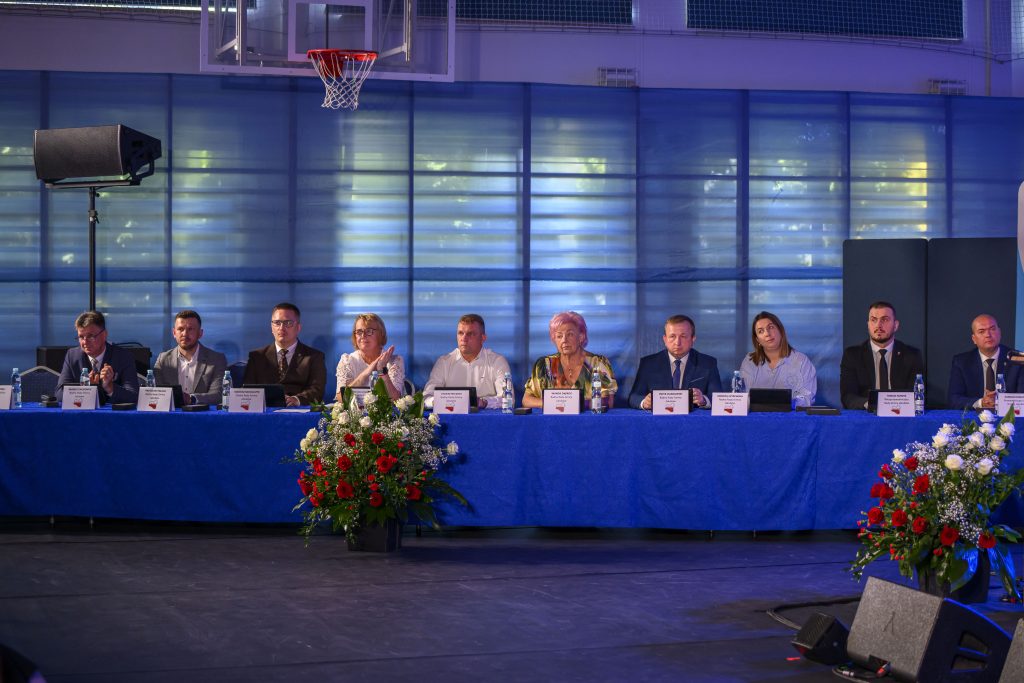 Grupa osób siedzi przy stole przykrytym niebieskim obrusem. Za nimi jest jasnoniebieskie, przeszklone tło. Przed stołem stoją bukiety kwiatów.