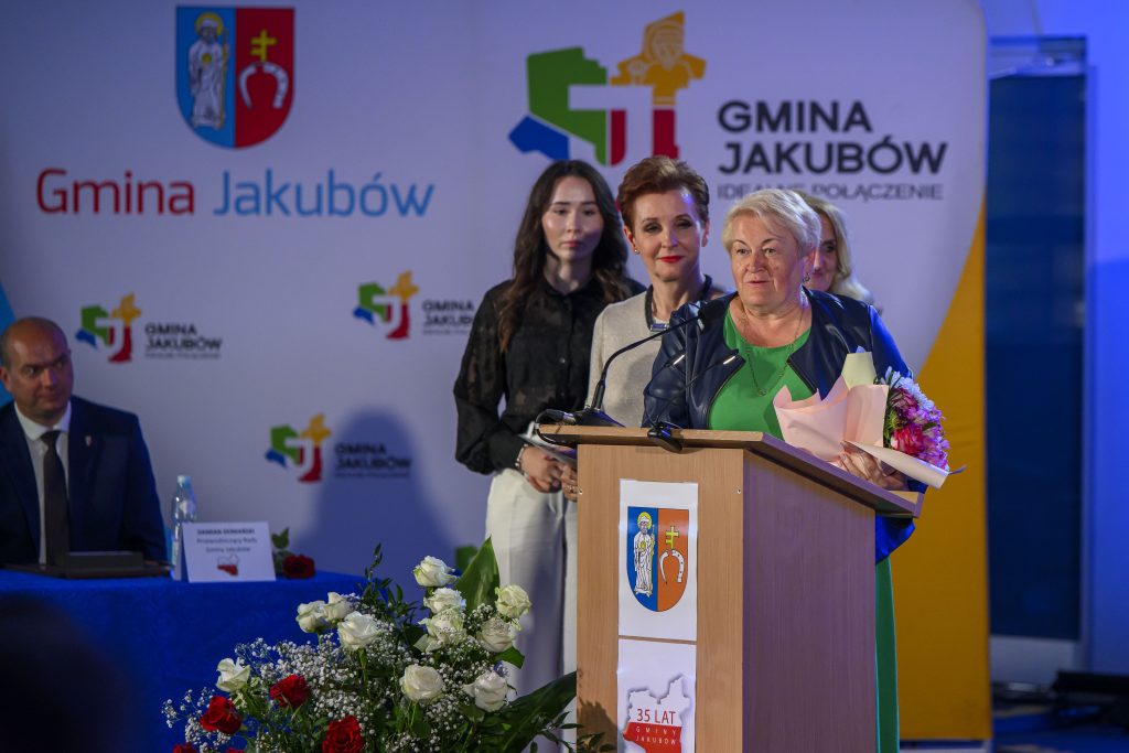 Kobieta z kwiatami przy podium, obok inne osoby. Tło z logotypem Gminy Jakubów i biało-czerwona dekoracja. Kwiaty w kolorach białym i czerwonym.