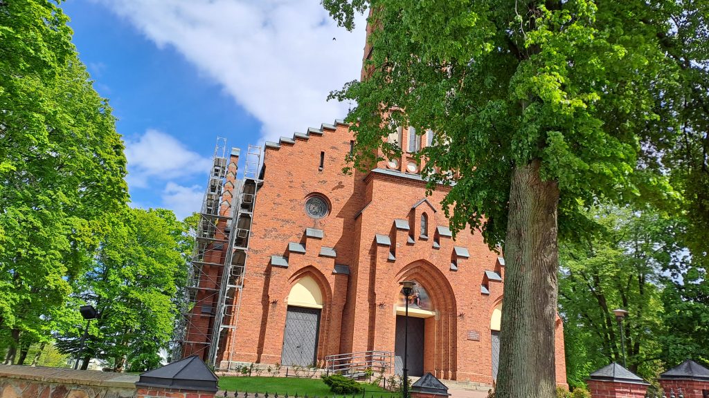 Ceglany kościół z rusztowaniem po lewej stronie. Drzewa otaczają budowlę, a niebo jest jasnoniebieskie z chmurami.
