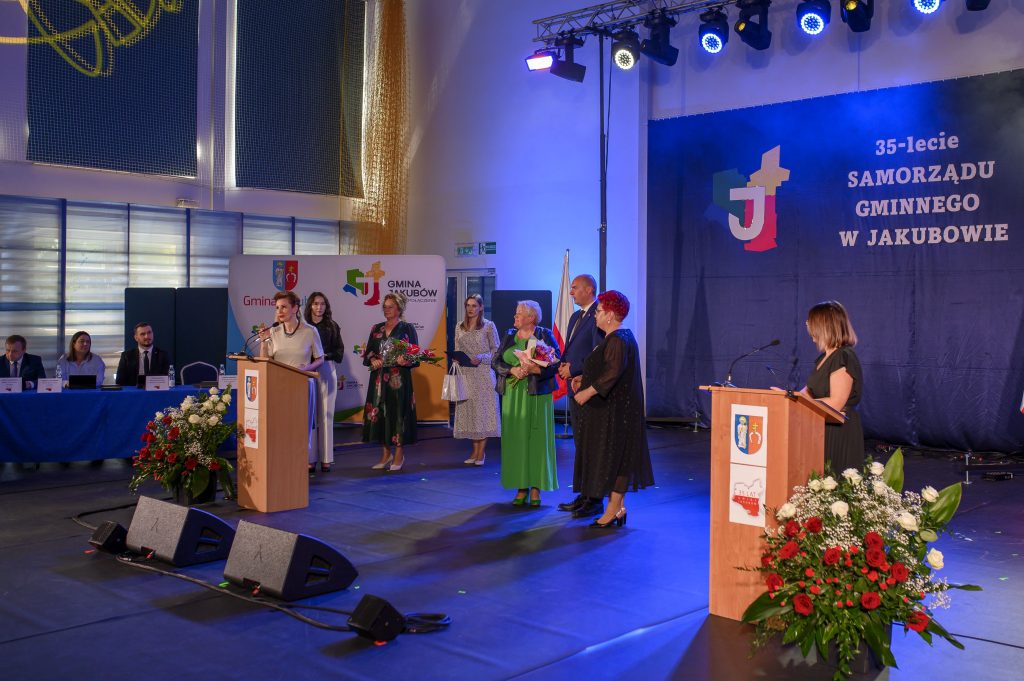 Grupa osób na scenie podczas uroczystości z okazji 35-lecia samorządu w Jakubowie. Dominują kolory niebieski i czerwony. Na tle ciemnoniebieskiej kurtyny znajduje się podium oraz dekoracje z kwiatami.