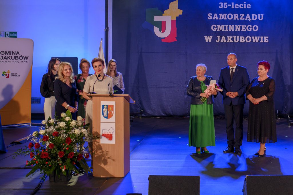 Grupa ludzi w eleganckich strojach stoi na scenie przy mikrofonie. W tle niebieski baner z napisem o 35-leciu samorządu w Jakubowie. Z przodu widoczne kwiaty w białej i czerwonej tonacji.