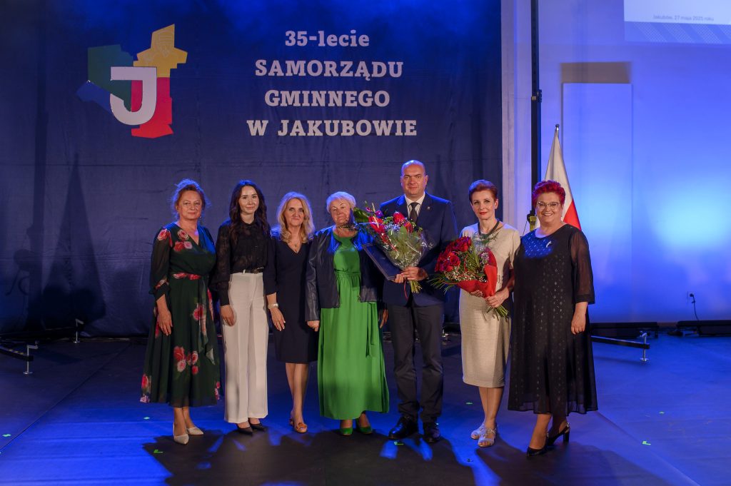 Na zdjęciu grupa ludzi pozuje na scenie z niebieskim tłem z napisem 35-lecie Samorządu Gminnego w Jakubowie. Mężczyzna w garniturze trzyma bukiet. Obok niego stoją kobiety w eleganckich strojach.