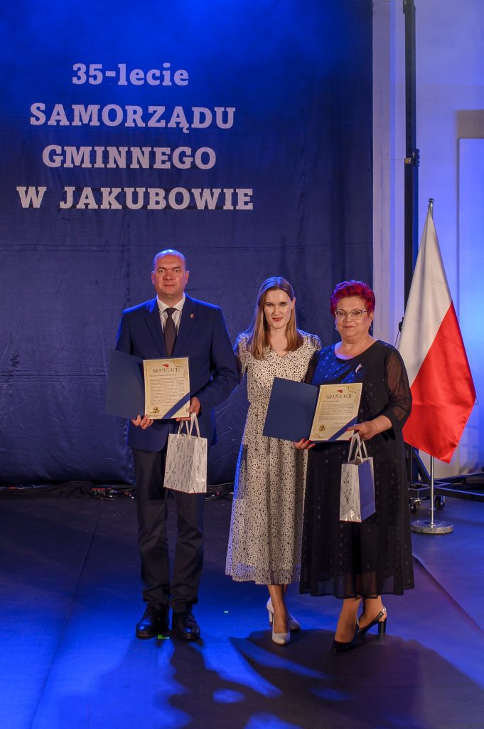 Trzy osoby stoją na tle granatowej kurtyny z napisem 35-lecie Samorządu Gminnego w Jakubowie. Każda trzyma certyfikat i torbę. Po prawej stronie widać polską flagę.