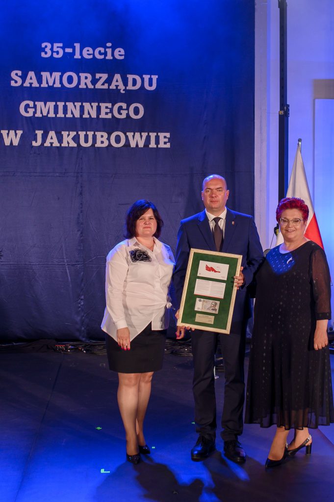 Trzy osoby stoją na scenie z granatowym tłem, trzymając dyplom. Kobiety w białej i czarnej sukience, mężczyzna w garniturze. Napis: 35-lecie Samorządu Gminnego w Jakubowie.