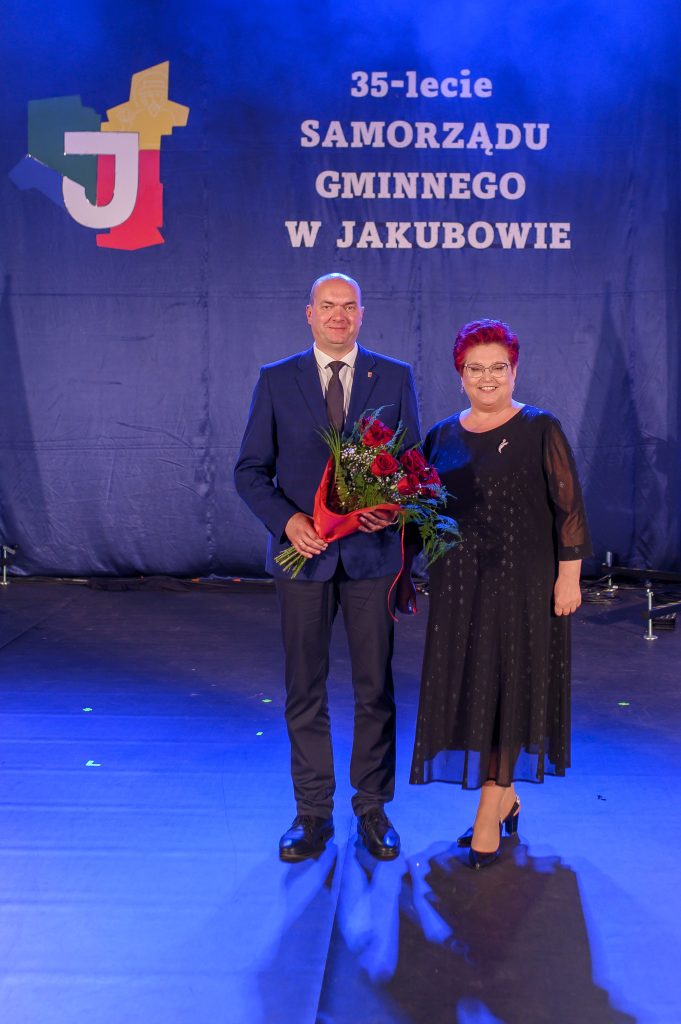 Dwójka ludzi na tle granatowej kurtyny; jeden trzyma bukiet kwiatów. Obok nich napis: 35-lecie samorządu gminnego w Jakubowie.