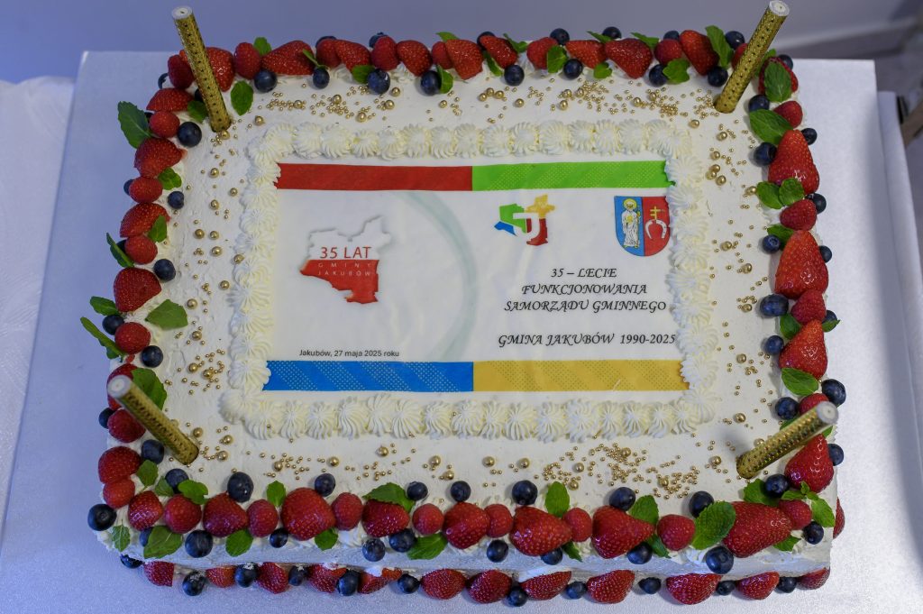 Prostokątny tort z owocami i kremem na białym tle. Na środku kolorowa grafika z mapą i tekstem o jubileuszu. Dekoracja z truskawek, borówek i złotych kuleczek.