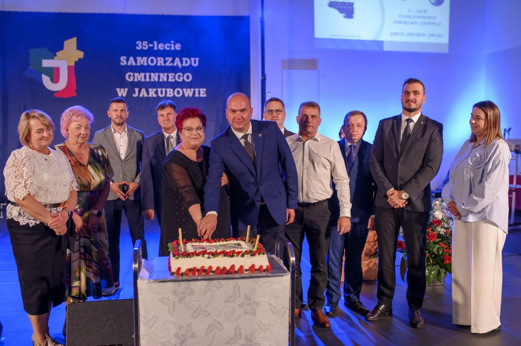Na tle niebieskiej sceny grupa osób przecina tort. Ubrania są eleganckie, w ciemnych i jasnych tonach. Obok znajduje się ozdobiony biały tort z owocami.
