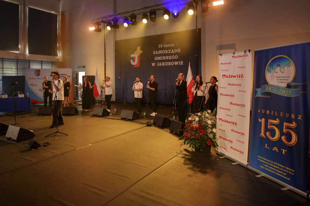 Grupa osób śpiewa na scenie przy mikrofonach. Tło czarne z napisem 35-lecie Samorządu Gminnego w Jakubowie. Po bokach plakaty i kwiaty.