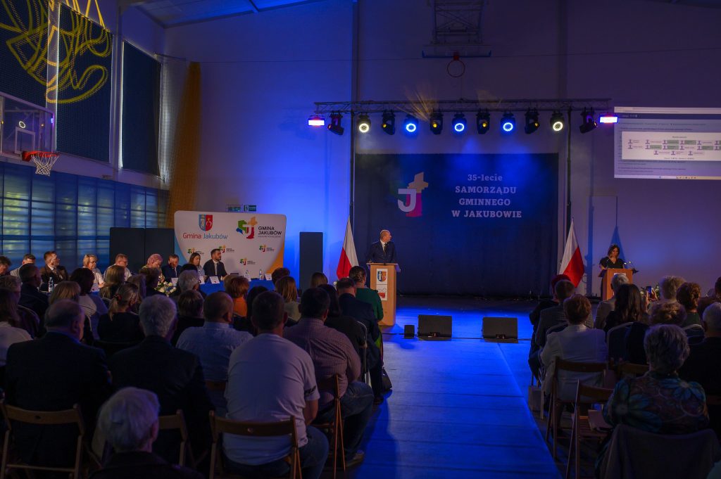 Na zdjęciu widoczne jest wydarzenie w hali, z tłumem ludzi siedzących i osoba przemawiająca. Dominują odcienie niebieskie i fioletowe z kolorowymi światłami na scenie. W tle znajdują się flagi oraz znak Gmina Jakubów.
