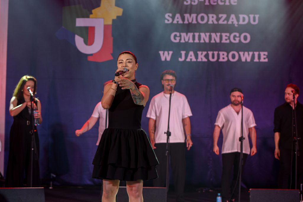 Na scenie występuje wokalistka w czarnej sukience, za nią chór. Tło to ciemnoniebieski baner z napisem 35-lecie Samorządu. Światła sceniczne oświetlają artystów.