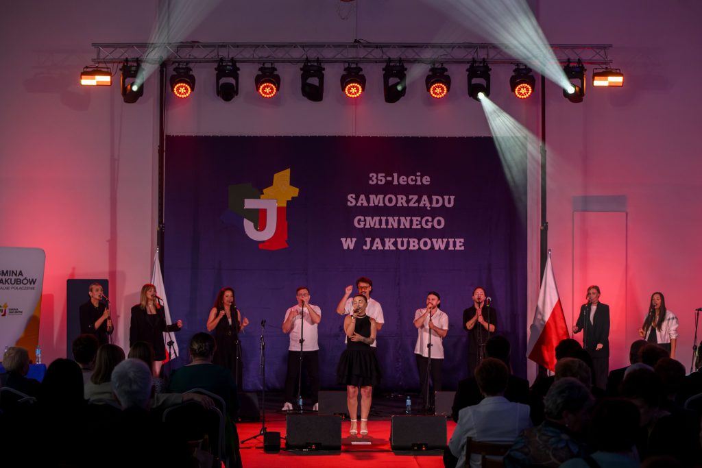 Na scenie oświetlonej reflektorami występuje zespół wokalny. Tło w kolorze fioletowym z napisem 35-lecie Samorządu Gminnego w Jakubowie. Po bokach widoczne flagi Polski.