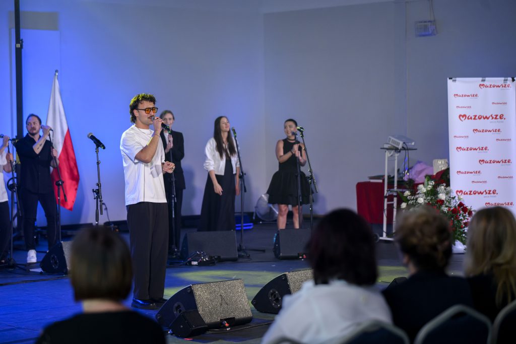 Grupa muzyków występuje na scenie, oświetlone niebieskim światłem. Przód sceny zajmuje mężczyzna w białej koszuli, trzymający mikrofon. W tle widoczny jest polski biało-czerwony banner.