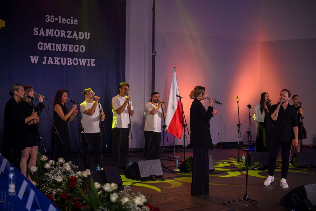 Grupa osób na scenie śpiewa przy mikrofonach. W tle napis 35-lecie Samorządu Gminnego w Jakubowie i flaga Polski. Scena oświetlona kolorowymi światłami.