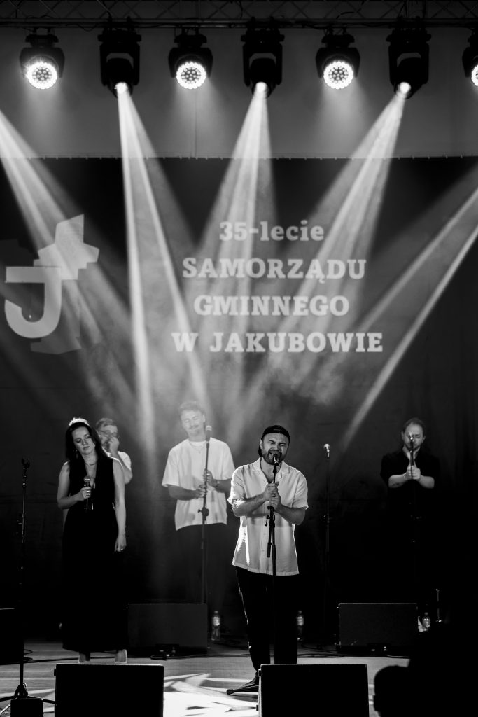 Ludzie śpiewają na scenie, tło z napisem 35-lecie Samorządu Gminnego w Jakubowie. Reflektory oświetlają scenę.