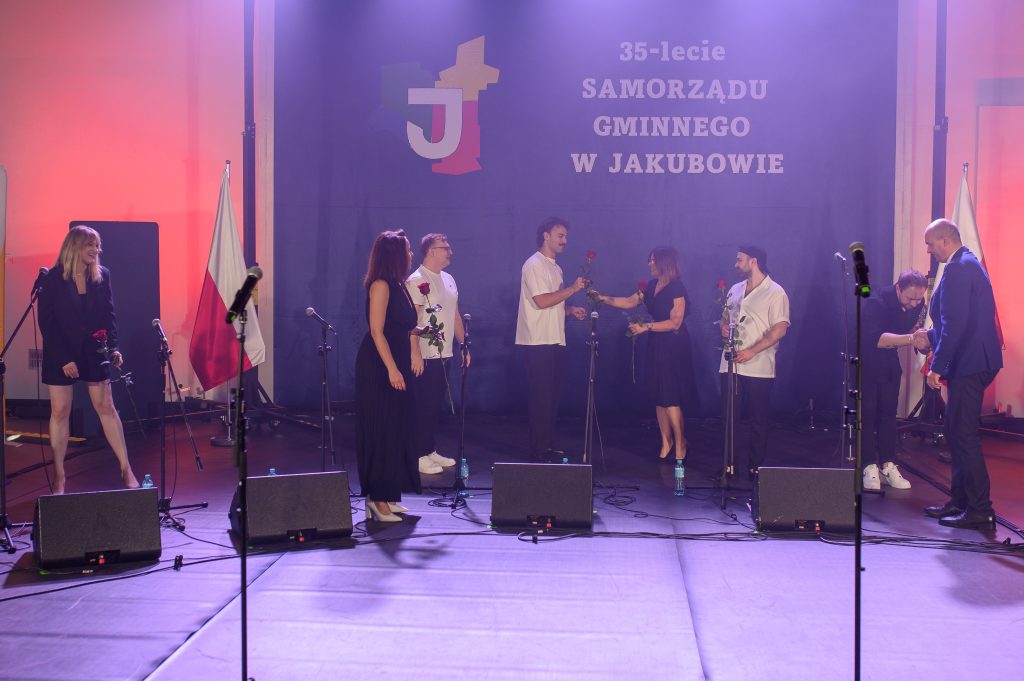 Grupa ludzi stoi na scenie z mikrofonami i różami, tło czarne z napisem 35-lecie Samorządu Gminnego w Jakubowie. Tło jest podświetlone różowymi światłami, a w tle widoczna jest polska flaga. Na scenie panuje
