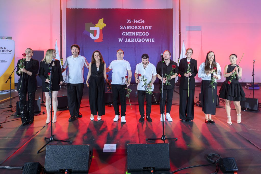 Grupa osób stoi na scenie pod napisem 35-lecie samorządu gminnego w Jakubowie. Mają na sobie eleganckie stroje i trzymają róże. Tło w odcieniach różu i fioletu.